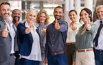 Diverse group of smiling business professionals giving thumbs up, standing together in an office setting.